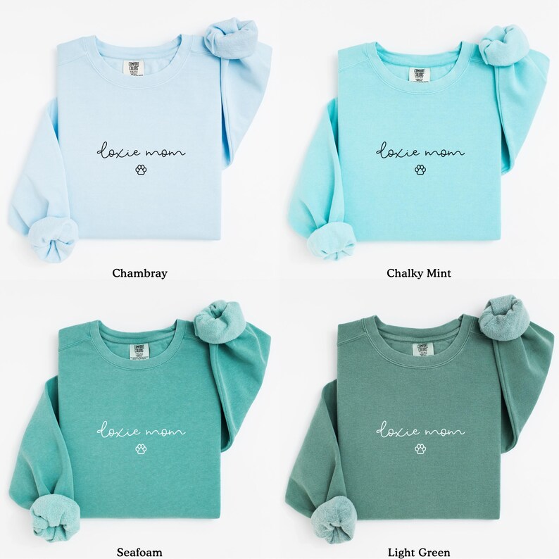 May include: Four long-sleeved sweatshirts in chambray, chalky mint, seafoam, and light green. Each shirt features the text "doxie mom" with a paw print graphic. The sleeves are rolled up, and the shirts are displayed on a white background.