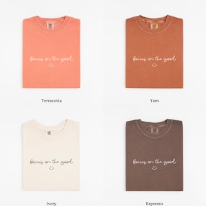 May include: Four folded t-shirts in terracotta, yam, ivory, and espresso colors. Each shirt has the phrase "focus on the good" and a smiling face printed on the front. The shirts are displayed on a white background.