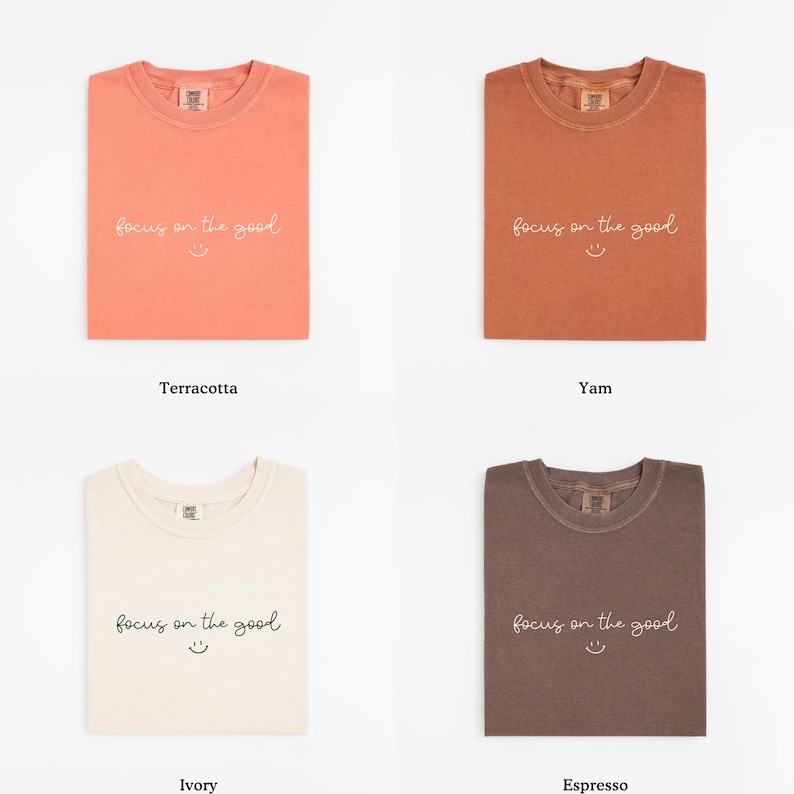 May include: Four folded t-shirts in terracotta, yam, ivory, and espresso colors. Each shirt has the phrase "focus on the good" and a smiling face printed on the front. The shirts are displayed on a white background.