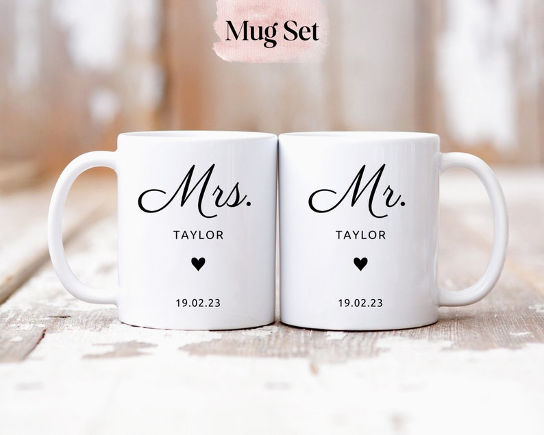 May include: A set of two white ceramic mugs with black lettering. The mugs say "Mrs. TAYLOR" and "Mr. TAYLOR" with a heart and the date "19.02.23" on each mug.