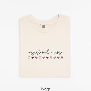Pode incluir: Camiseta cor marfim com as palavras "registered nurse" em fonte manuscrita, com cora&ccedil;&otilde;es rosa por baixo. A camisa est&aacute; dobrada cuidadosamente, e a etiqueta da marca &eacute; vis&iacute;vel no topo. A palavra "Ivory" est&aacute; impressa na parte inferior.