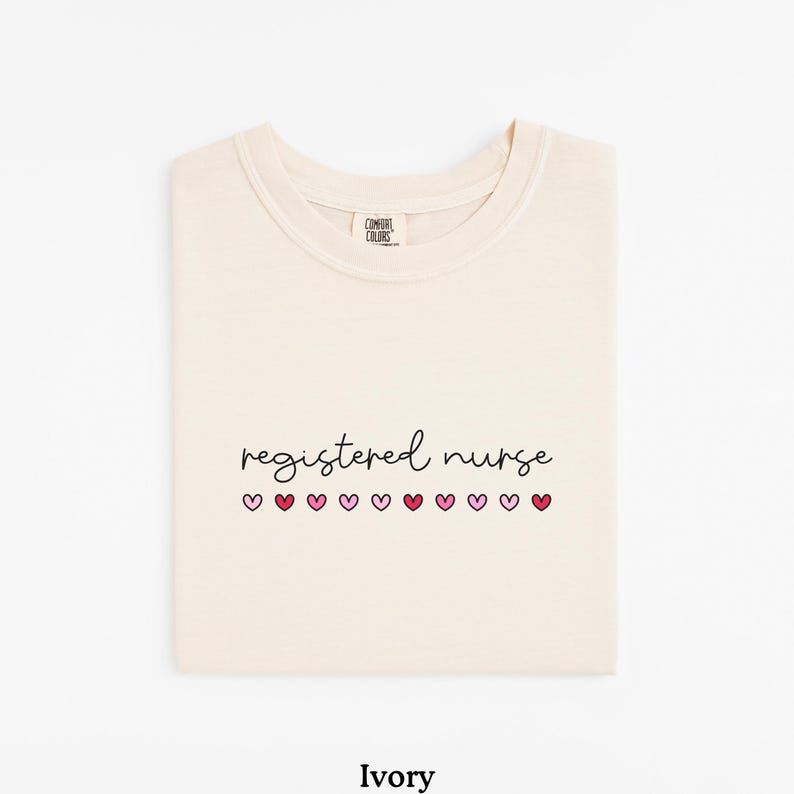 Pode incluir: Camiseta cor marfim com as palavras "registered nurse" em fonte manuscrita, com cora&ccedil;&otilde;es rosa por baixo. A camisa est&aacute; dobrada cuidadosamente, e a etiqueta da marca &eacute; vis&iacute;vel no topo. A palavra "Ivory" est&aacute; impressa na parte inferior.