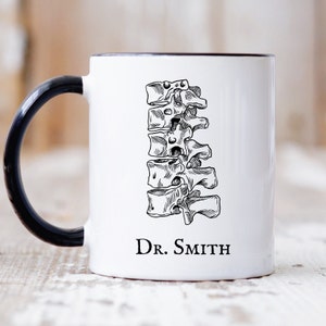 May include: White ceramic coffee mug with a black handle. The mug features a black and white illustration of a human spine and the text "Dr. Smith".
