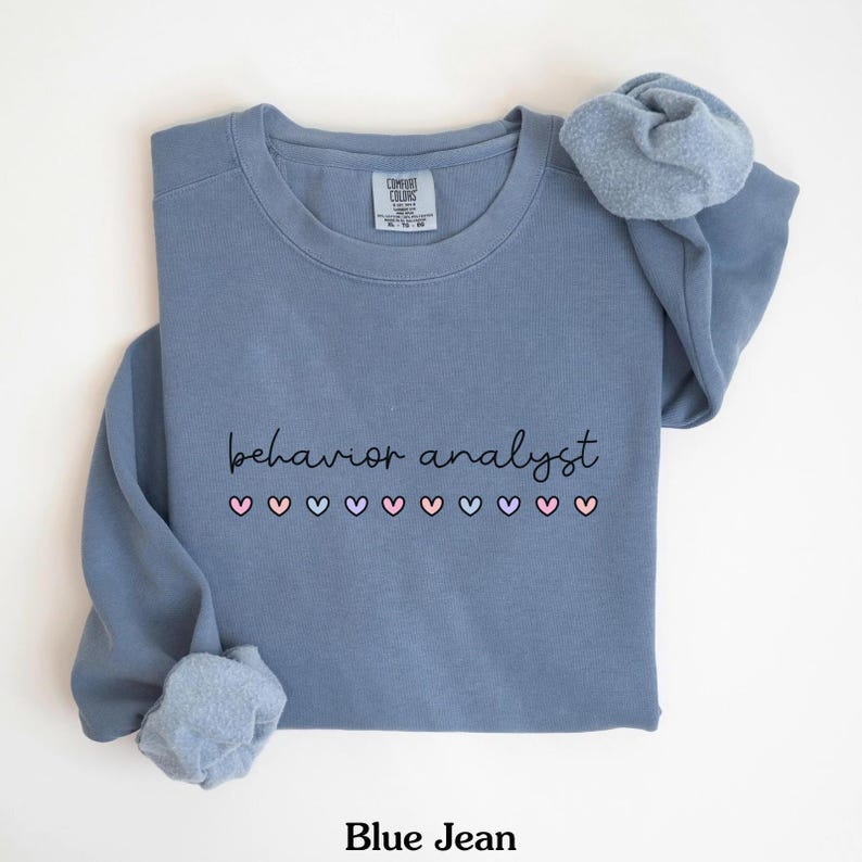 K&ouml;nnte beinhalten: Ein blaues Sweatshirt mit dem Text "behavior analyst" und einer Reihe von rosa, blauen und wei&szlig;en Herzen.