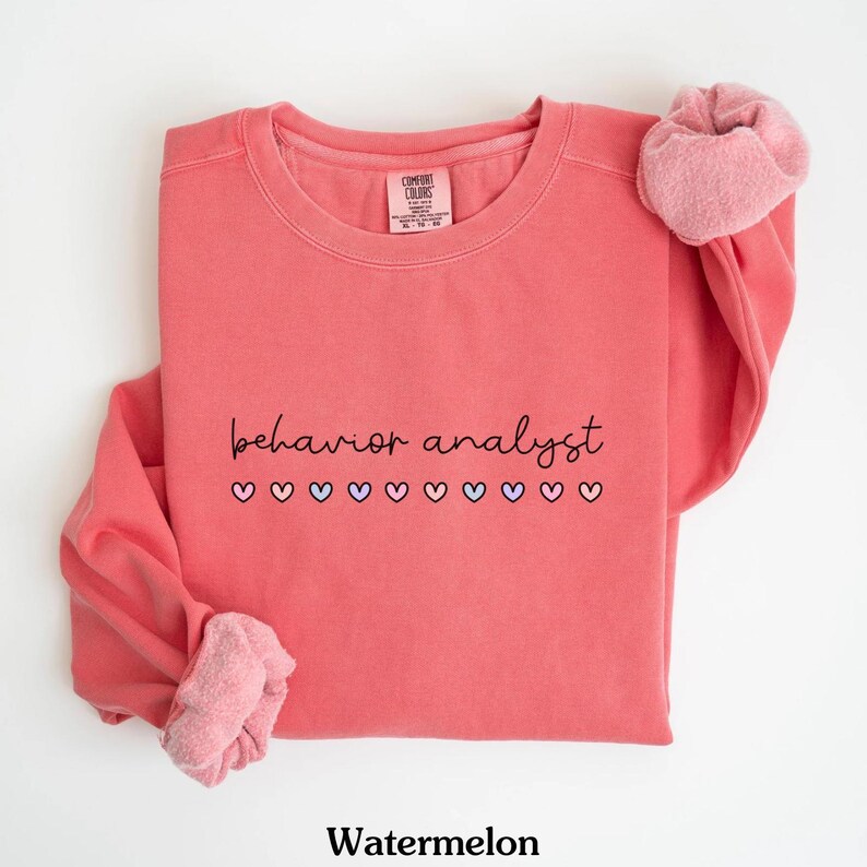 K&ouml;nnte beinhalten: Wassermelonenfarbener Pullover mit dem Text "behavior analyst" in schwarzer Schrift und einer Reihe kleiner rosa, lila und blauer Herzen darunter.