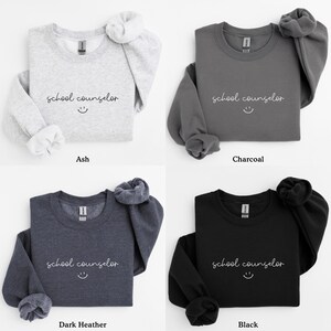 Pode incluir: Quatro sweatshirts de gola redonda em diferentes cores: cinza, carv&atilde;o, cinza escuro e preto. Cada sweatshirt apresenta o texto "school counselor" com um rosto sorridente.