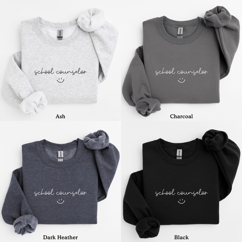 Pode incluir: Quatro sweatshirts de gola redonda em diferentes cores: cinza, carv&atilde;o, cinza escuro e preto. Cada sweatshirt apresenta o texto "school counselor" com um rosto sorridente.