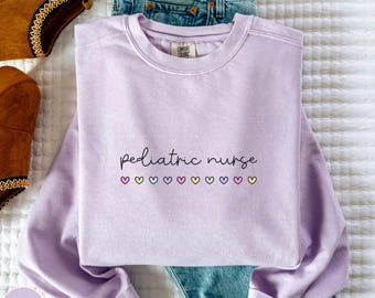 Puede incluir: Sudadera lila con la frase "pediatric nurse" en escritura negra. Debajo, varios corazones pequeños y coloridos. La sudadera está doblada sobre unos vaqueros azules y junto a unas botas marrones.