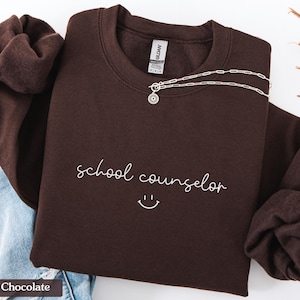 Pode incluir: Camisola de cor castanho chocolate escuro com o texto "orientador escolar" e um sorriso branco.