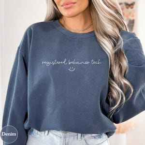 May include: A denim blue sweatshirt with the text "registered behavior tech" in a cursive font, and a small smiling face. The sweatshirt has a crew neck and long sleeves. The word "Denim" is printed on the bottom left corner.