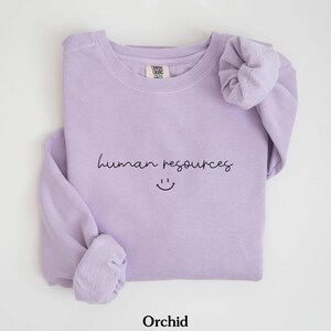 May include: A lavender sweatshirt with the text "human resources" and a smiley face. The sweatshirt is folded and has a scrunchie on the sleeve.