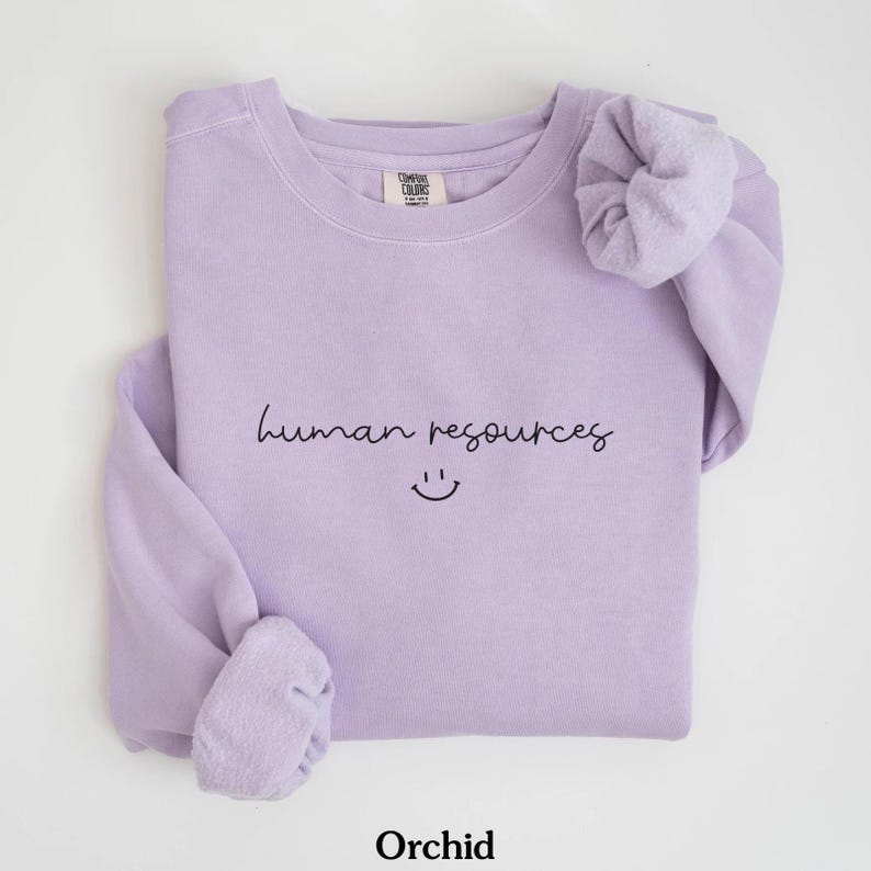 May include: A lavender sweatshirt with the text "human resources" and a smiley face. The sweatshirt is folded and has a scrunchie on the sleeve.