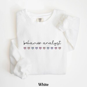 K&ouml;nnte beinhalten: Wei&szlig;er Pullover mit dem Text "behavior analyst" und einer Reihe von rosa, blauen und violetten Herzen.