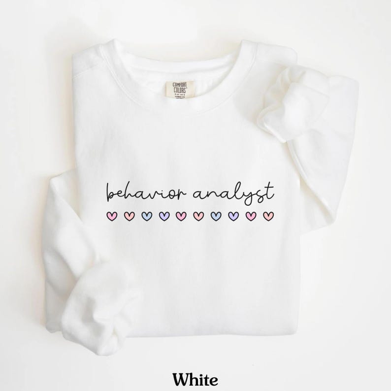 K&ouml;nnte beinhalten: Wei&szlig;er Pullover mit dem Text "behavior analyst" und einer Reihe von rosa, blauen und violetten Herzen.