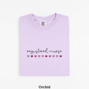 Pode incluir: Camisola de cor orqu&iacute;dea com as palavras "registered nurse" em escrita preta, com cora&ccedil;&otilde;es rosa por baixo. A camisola est&aacute; dobrada cuidadosamente, com a etiqueta da marca vis&iacute;vel na gola.