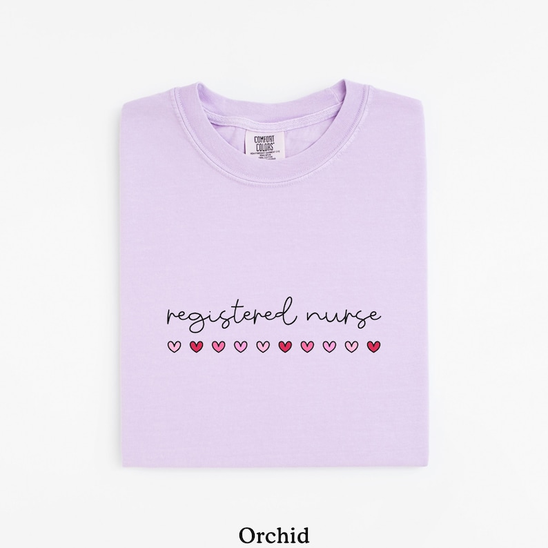 Pode incluir: Camisola de cor orqu&iacute;dea com as palavras "registered nurse" em escrita preta, com cora&ccedil;&otilde;es rosa por baixo. A camisola est&aacute; dobrada cuidadosamente, com a etiqueta da marca vis&iacute;vel na gola.