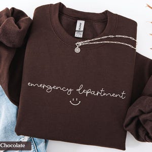 May include: Dark chocolate brown sweatshirt with the text "emergency department" and a smiley face in white.
