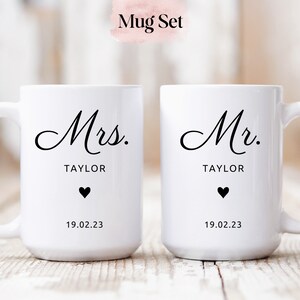 May include: A set of two white ceramic mugs with black text. The mug on the left says "Mrs. TAYLOR" with a small black heart and the date "19.02.23". The mug on the right says "Mr. TAYLOR" with a small black heart and the date "19.02.23".