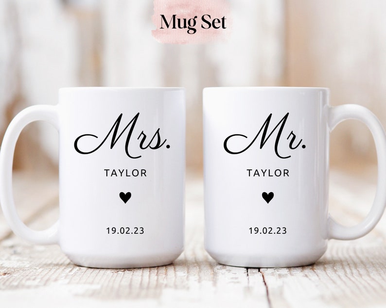 May include: A set of two white ceramic mugs with black text. The mug on the left says "Mrs. TAYLOR" with a small black heart and the date "19.02.23". The mug on the right says "Mr. TAYLOR" with a small black heart and the date "19.02.23".