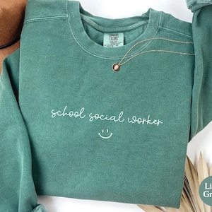 Puede incluir: Una sudadera verde claro con el texto "school social worker" y una carita sonriente en blanco.