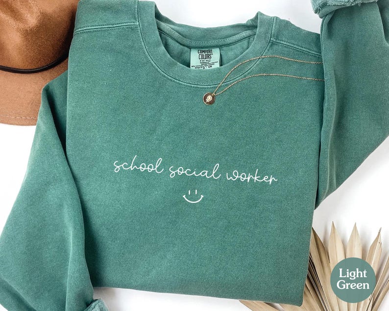 Puede incluir: Una sudadera verde claro con el texto "school social worker" y una carita sonriente en blanco.