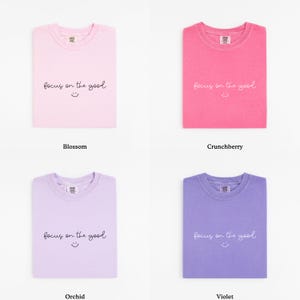 May include: Four folded t-shirts in different colors: blossom, crunchberry, orchid, and violet. Each shirt has the phrase "focus on the good" and a smiley face printed on the front.
