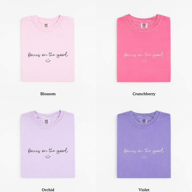 May include: Four folded t-shirts in different colors: blossom, crunchberry, orchid, and violet. Each shirt has the phrase "focus on the good" and a smiley face printed on the front.