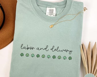 May include: A light green Comfort Colors t-shirt with the words "labor and delivery" in black script, and green shamrocks. A gold necklace with a small pendant rests on the shirt. The shirt is folded.