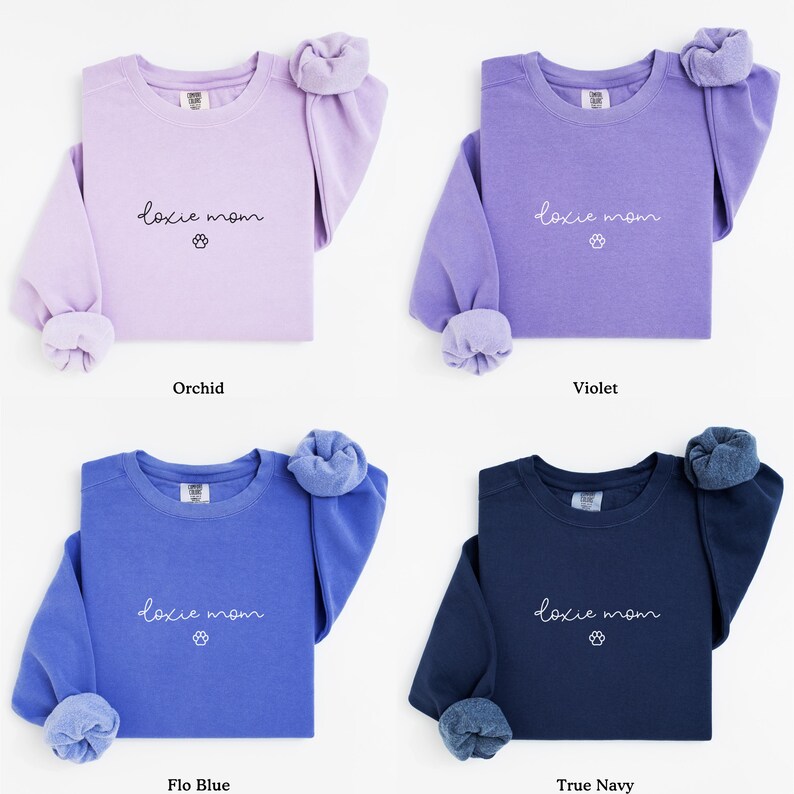 May include: Four crewneck sweatshirts in orchid, violet, flo blue, and true navy. Each sweatshirt has the text "doxie mom" with a paw print graphic. The sleeves are cuffed, and the colors are solid.