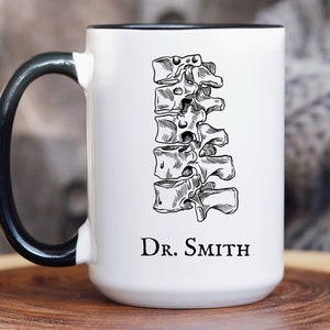 May include: White ceramic mug with a black handle. The mug features a black and white illustration of a human spine and the text "Dr. Smith".