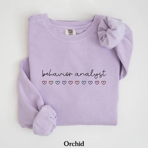 K&ouml;nnte beinhalten: Ein lavendelfarbener Pullover mit dem Text "behavior analyst" in schwarzer Schrift und einer Reihe bunter Herzen darunter. Der Pullover hat einen Rundhalsausschnitt und lange &Auml;rmel.