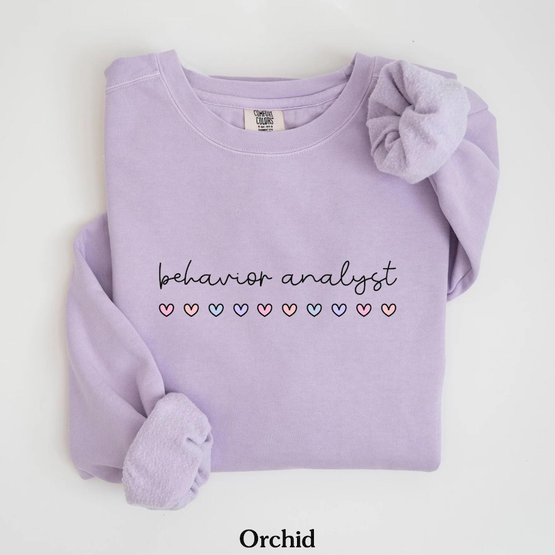 K&ouml;nnte beinhalten: Ein lavendelfarbener Pullover mit dem Text "behavior analyst" in schwarzer Schrift und einer Reihe bunter Herzen darunter. Der Pullover hat einen Rundhalsausschnitt und lange &Auml;rmel.