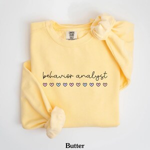K&ouml;nnte beinhalten: Ein gelber Pullover mit dem Text "behavior analyst" in schwarzer Schrift und einer Reihe von rosa, blauen und wei&szlig;en Herzen. Der Pullover ist von Comfort Colors.
