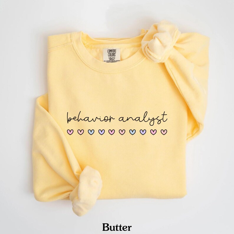 K&ouml;nnte beinhalten: Ein gelber Pullover mit dem Text "behavior analyst" in schwarzer Schrift und einer Reihe von rosa, blauen und wei&szlig;en Herzen. Der Pullover ist von Comfort Colors.