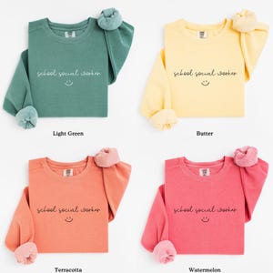 Puede incluir: Cuatro sudaderas de manga larga en diferentes colores: verde claro, amarillo, naranja y rosa. Cada sudadera tiene el texto "school social worker" con una carita sonriente impresa en la parte delantera.