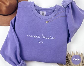 May include: A violet-colored sweatshirt with the words "music teacher" in white script and a smiling face. A gold necklace with a pendant is draped on the sweatshirt. A brown hat is visible in the upper left corner.