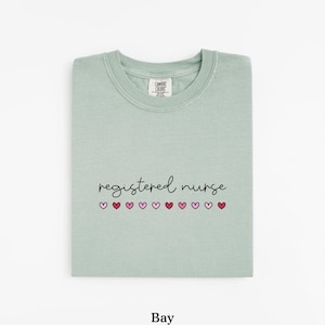 Pode incluir: Camisola verde-claro com as palavras "registered nurse" em escrita preta, com cora&ccedil;&otilde;es rosa por baixo. A palavra "Bay" est&aacute; impressa na parte inferior da camisola.