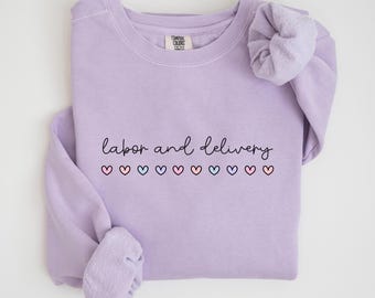 Könnte beinhalten: Ein lavendelfarbener Pullover mit dem Text "Arbeit und Geburt" in schwarzer Schrift. Unter dem Text befindet sich eine Reihe kleiner, bunter Herzen.