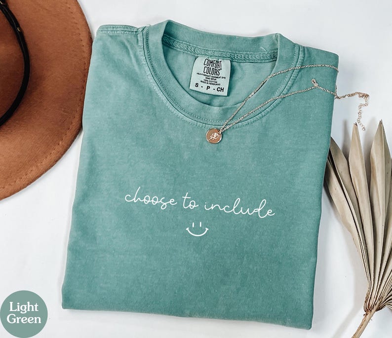 May include: A light green t-shirt with the phrase "choose to include" and a smiley face printed in white. A gold necklace with a pendant is draped across the shirt.