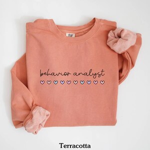 K&ouml;nnte beinhalten: Terracottafarbener Pullover mit dem Text "behavior analyst" in schwarzer Schrift und einer Reihe von rosa, blauen und violetten Herzen.