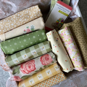 May include: A collection of rolled fabric bundles in a white box, featuring various floral and patterned designs in shades of yellow, green, and pink. Includes a pair of green scissors and a charm package labeled "Sew Charming Dresden Enamel Charm."