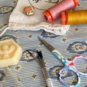 May include: A collection of sewing supplies including a small bag with the text "Tiny Stitch", two spools of thread, one pink and one yellow, a small container of sewing needles, and a pair of colorful scissors.