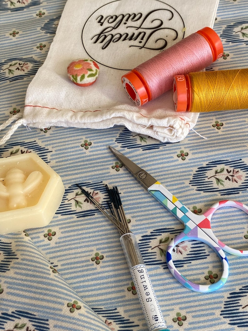 May include: A collection of sewing supplies including a small bag with the text "Tiny Stitch", two spools of thread, one pink and one yellow, a small container of sewing needles, and a pair of colorful scissors.
