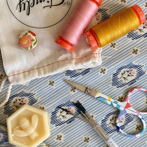 May include: A close-up of a sewing kit with a variety of tools, including a small bag, two spools of thread, a small button, a hexagonal block of beeswax, a pair of scissors, and a pack of needles. The fabric background is blue and white with a floral pattern.