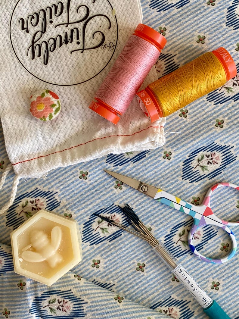May include: A close-up of a sewing kit with a variety of tools, including a small bag, two spools of thread, a small button, a hexagonal block of beeswax, a pair of scissors, and a pack of needles. The fabric background is blue and white with a floral pattern.