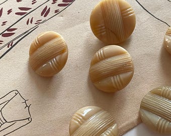 Puede incluir: Una colección de botones redondos vintage en tonos beige y crema. Cada botón presenta un diseño a rayas y entrecruzado. Los botones están dispuestos sobre un fondo de papel marrón claro, lo que sugiere un tema de costura o artesanía.