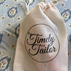 May include: A white cotton drawstring bag with the text "the Timely Tailor" printed in black on a circle.
