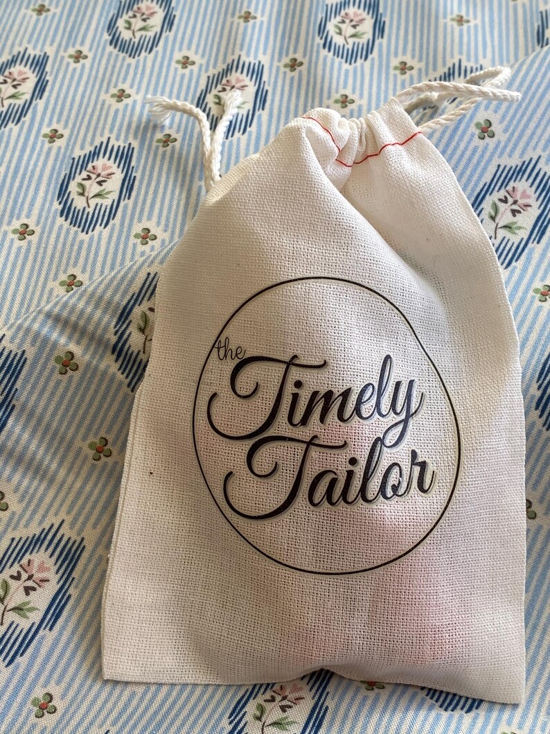 May include: A white cotton drawstring bag with the text "the Timely Tailor" printed in black on a circle.