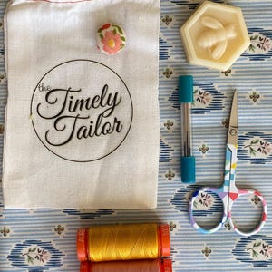 May include: A white cotton drawstring bag with the text "the Timely Tailor" printed on it. The bag is next to a small floral button, a hexagonal soap bar with a bee design, a turquoise and silver needle holder, a pair of colorful scissors, and two spools of thread, one pink and one yellow.