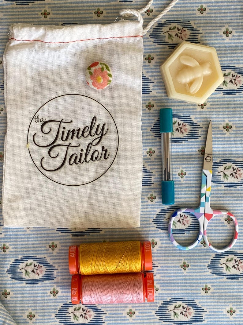 May include: A white cotton drawstring bag with the text "the Timely Tailor" printed on it. The bag is next to a small floral button, a hexagonal soap bar with a bee design, a turquoise and silver needle holder, a pair of colorful scissors, and two spools of thread, one pink and one yellow.